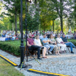 Zdjęcie przedstawia grupę ludzi siedzących na krzesłach na zewnątrz w parku. Tło stanowią zielone drzewa i trawnik. Widoczne są też stoły z białymi obrusami.