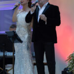 Dwoje ludzi śpiewa na scenie, ona w białej sukni, on w ciemnym garniturze. Tło jasne z lampionem. Klawiatura w tle.