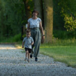 Kobieta i chłopiec spacerują żwirową ścieżką w parku. Wokół widać zieleń drzew. Ubrania w stonowanych kolorach, tło jest zielone i spokojne.