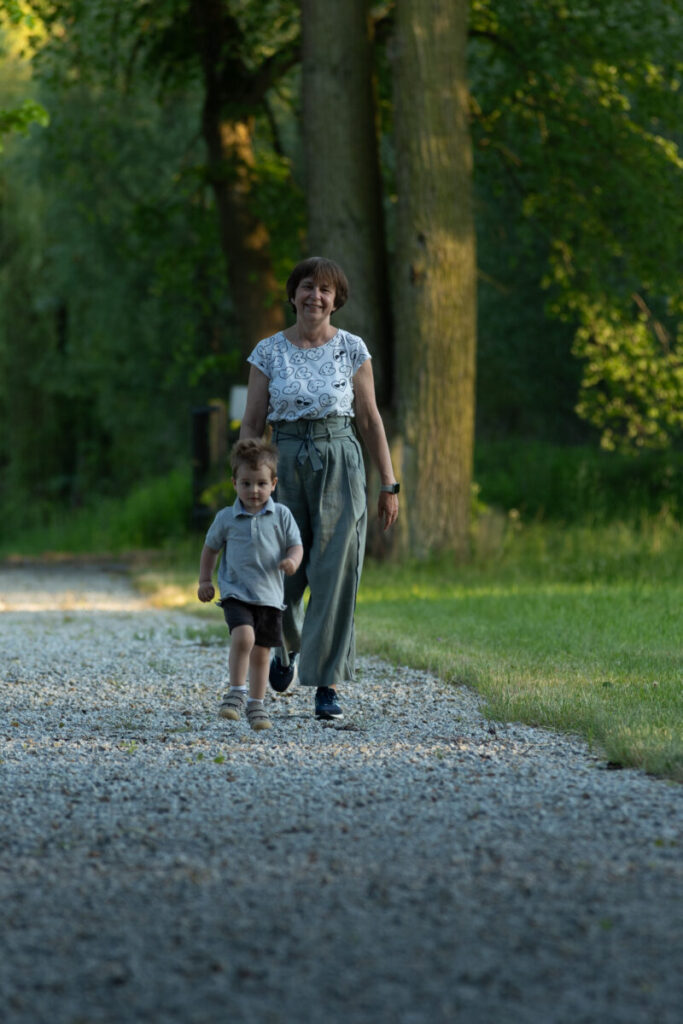 Kobieta i chłopiec spacerują żwirową ścieżką w parku. Wokół widać zieleń drzew. Ubrania w stonowanych kolorach, tło jest zielone i spokojne.
