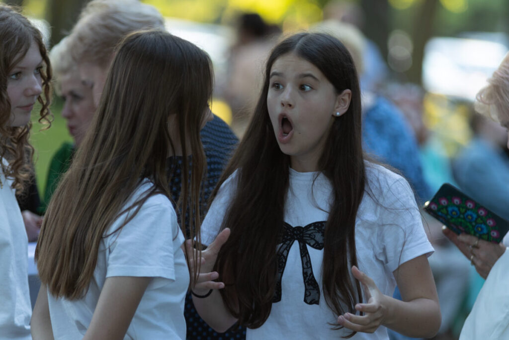 Na zdjęciu są dwie dziewczyny w białych koszulkach; jedna z rozdziawionymi ustami. Tło jest rozmyte, pełne zieleni. Inne osoby są w tle.