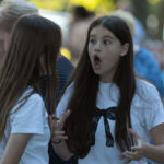 Na zdjęciu są dwie dziewczyny w białych koszulkach; jedna z rozdziawionymi ustami. Tło jest rozmyte, pełne zieleni. Inne osoby są w tle.