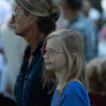 Na zdjęciu widoczna jest kobieta i dziecko w okularach. Osoby są w tłumie; kobieta ma ciemny strój, a dziecko niebieski. Tło jest rozmyte, dominują jasne kolory.