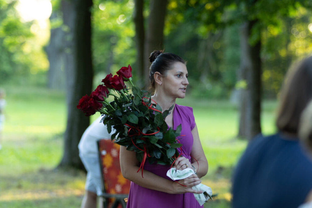 Kobieta w różowej sukience trzyma bukiet czerwonych róż. Tło to zielony las z promieniami słońca. Scena jest letnia i spokojna.