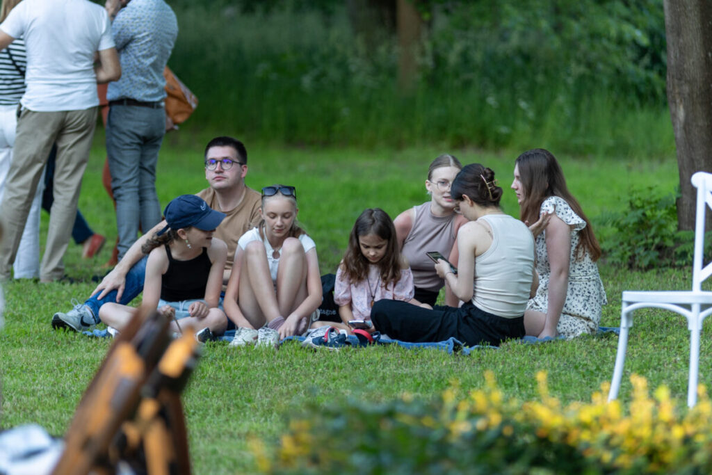 Na zdjęciu grupa młodych ludzi siedzi na trawie w parku. Ubrania są w jasnych odcieniach, a tło to zielona roślinność. Jedna osoba trzyma telefon, reszta rozmawia.