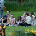 Na zdjęciu grupa młodych ludzi siedzi na trawie w parku. Ubrania są w jasnych odcieniach, a tło to zielona roślinność. Jedna osoba trzyma telefon, reszta rozmawia.