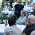 Na zdjęciu widać ludzi siedzących przy białych stołach na przyjęciu. Na stołach zielone dekoracje z liści. W tle inni goście i ogród.