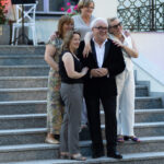 Pięć osób pozuje na marmurowych schodach. Tło zdobi krata i rośliny. Grupa elegancko ubrana, śmieje się i obejmuje.