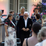 Grupa ludzi na schodach przed budynkiem. Kobiety w kolorowych strojach i mężczyzna w ciemnej marynarce. Tło z roślinnością i oświetleniem.