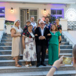 Grupa ludzi pozuje na schodach przed budynkiem z kolorowym oświetleniem. Zdjęcie robione jest z dołu, widać telefon w prawym dolnym rogu. Tło ma jasne ściany i fioletowe akcenty świetlne.