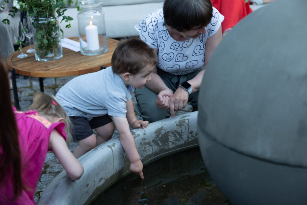 Dzieci bawią się przy fontannie, zanurzając ręce w wodzie. Kobieta w białej bluzce z wzorem serc obserwuje, siedząc obok. Stół z kwiatami i świecą w tle.