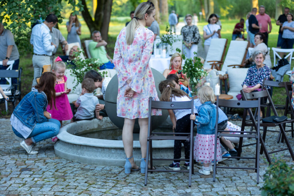 Grupa ludzi na zewnątrz przy fontannie. Dzieci bawią się wodą, otoczone dorosłymi. Tło to drzewa i zielona przestrzeń.