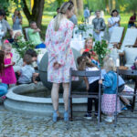 Grupa ludzi na zewnątrz przy fontannie. Dzieci bawią się wodą, otoczone dorosłymi. Tło to drzewa i zielona przestrzeń.