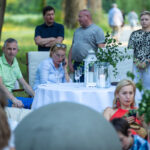 Ludzie siedzą przy białym stole z dekoracjami, świece, zielone rośliny. W tle drzewa i trawnik. Atmosfera pikniku.