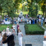 Obraz przedstawia tłum ludzi na zewnątrz, na brukowanym placu, otoczonym zielenią. Stoły z białymi obrusami i krzesłami są rozstawione wokół. W tle widać drzewa i prześwitujące światło słoneczne.