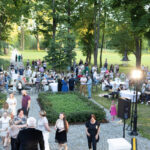 Na zdjęciu widoczny jest tłum ludzi na zewnątrz podczas eventu w parku. W tle drzewa i zielona trawa, na pierwszym planie stoły i krzesła. Dominuje zieleń i delikatne kontrasty bieli.