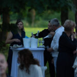 Grupa gości przy stole na zewnątrz w parku, serwująca napoje. W tle drzewa i zielony trawnik. Eleganckie ubrania, stół biały.