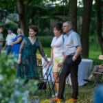 Grupa ludzi stoi na trawie w ogrodzie. Tła z drzewami i budynkiem. Osoby w letnich ubraniach, skupione rozmowach.