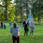Zdjęcie ukazuje ludzi spacerujących po trawie w parku. W tle widać pomnik otoczony drzewami. Dominuje zieleń trawy i liści.