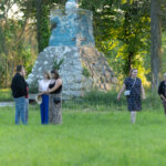 Grupa ludzi stoi na trawie przed kamiennym postumentem. W tle drzewa i zielona roślinność. Scena oświetlona popołudniowym słońcem.