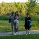 Trzy kobiety spacerują po żwirowej ścieżce w parku. Mają na sobie jasne ubrania, a w tle widać zieleń drzew. Światło jest miękkie, tworzy ciepłą atmosferę.