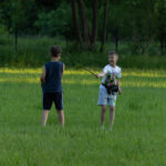 Dwóch chłopców bawi się na zielonej łące; jeden z nich trzyma patyk. W tle drzewa i krzewy. Dzień jest słoneczny.