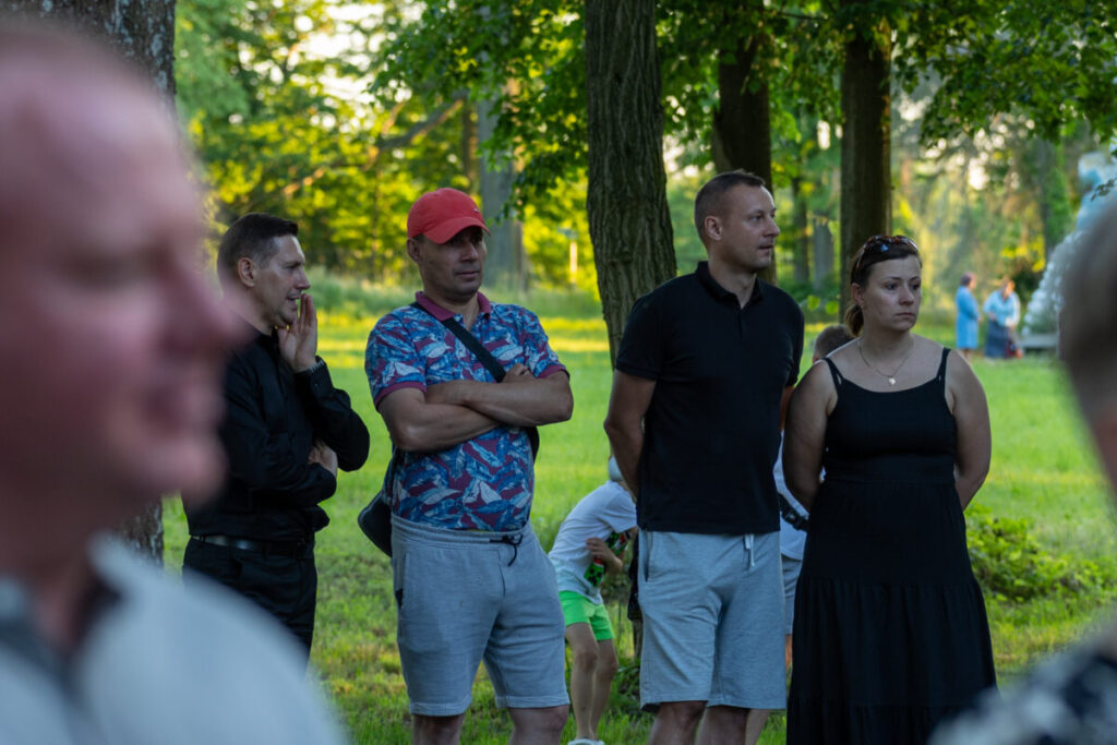 Cztery osoby stoją w parku, dwie z nich z założonymi rękami. Jeden mężczyzna w czerwonej czapce, kobieta w czarnej sukience. Tło to zielone drzewa i trawa.
