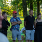 Cztery osoby stoją w parku, dwie z nich z założonymi rękami. Jeden mężczyzna w czerwonej czapce, kobieta w czarnej sukience. Tło to zielone drzewa i trawa.