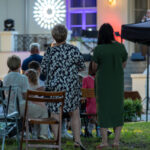 Grupa ludzi siedzi na zewnątrz, przed budynkiem z jasnymi ścianami. W tle scena z kolorowym oświetleniem. Wokół rośliny i namiot ogrodowy.