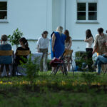 Zdjęcie przedstawia grupę ludzi na trawniku przed białym budynkiem. Stoją i siedzą przy stole z białym obrusem. W tle są drzewa i krzesła ogrodowe.
