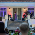 Scena plenerowa z artystką w fioletowej sukni na środku, otoczona muzykami. Tło to jasny budynek z kolorowymi światłami. Publiczność siedzi przed sceną na trawie.