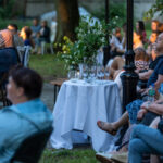 Na zdjęciu ludzie siedzą na zewnątrz, wokół stołu z białym obrusem i kieliszkami. Tło to zielone drzewa i lekko oświetlona scena. Atmosfera jest spokojna, wieczorna.