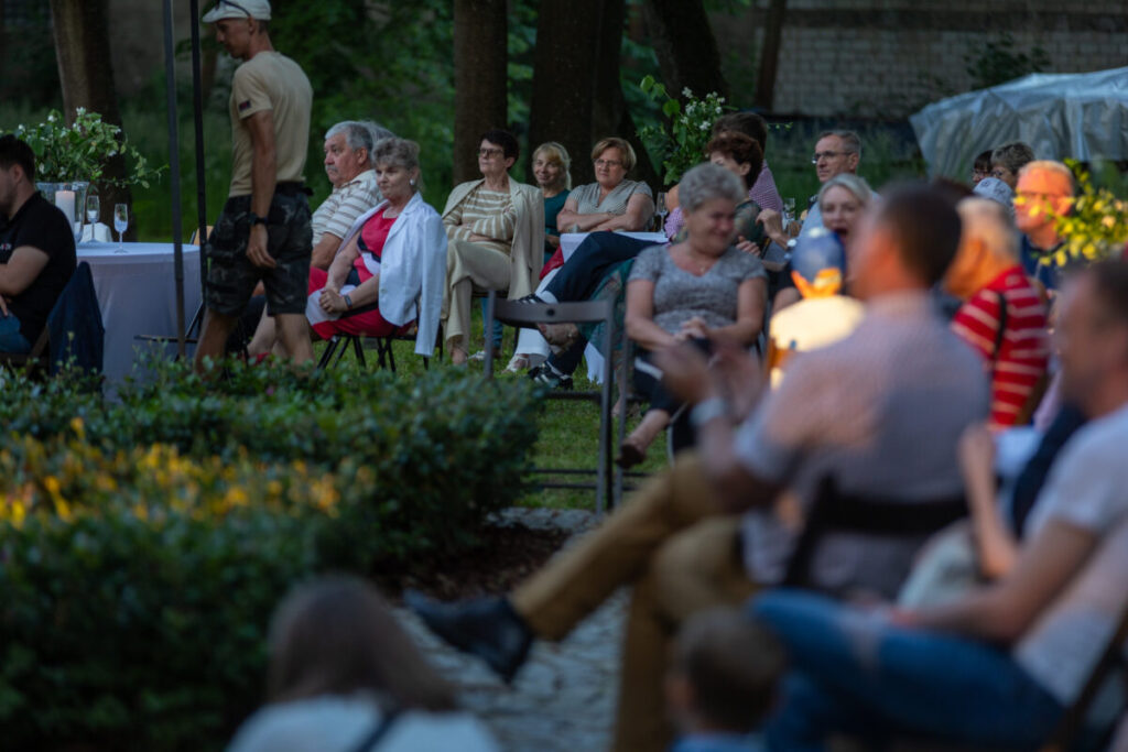 Grupa osób siedzi na zewnątrz przy stołach, wokół roślin. Tło zielone, wieczorne światło dodaje przytulności. Scena jest spokojna i towarzyska.