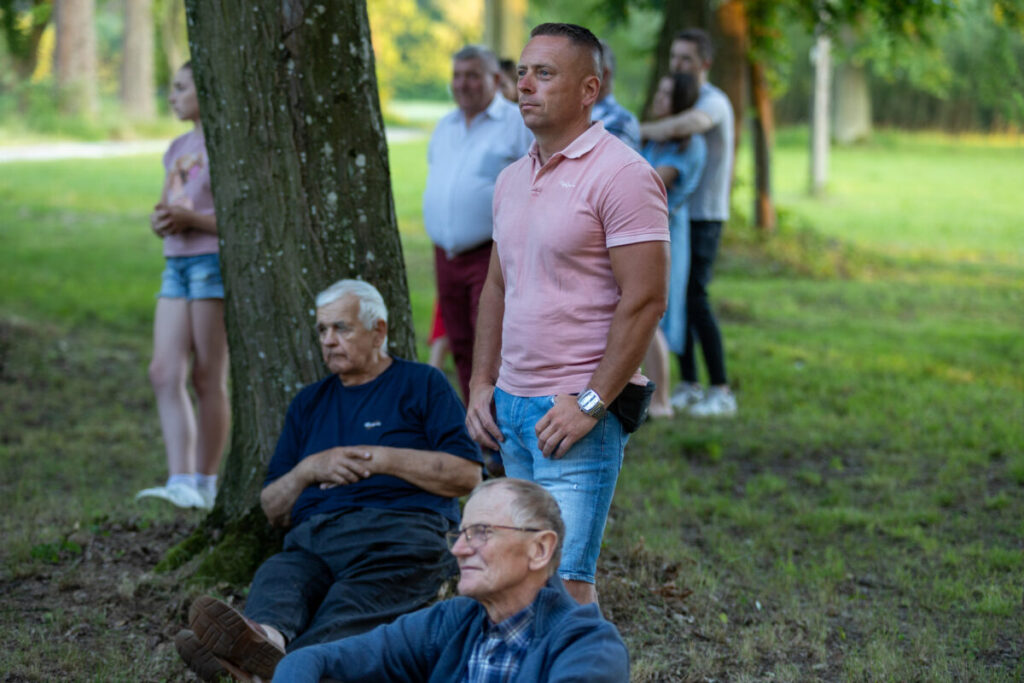 Mężczyźni siedzą przy drzewach na zielonej trawie. Jeden z nich stoi w różowej koszulce. W tle inne osoby i drzewa.