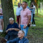 Mężczyźni siedzą przy drzewach na zielonej trawie. Jeden z nich stoi w różowej koszulce. W tle inne osoby i drzewa.