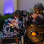 Dwóch mężczyzn na wózkach, jeden z telefonem. Ubrani kolorowo, w tle niebiesko podświetlona ściana. Atmosfera wieczorna.