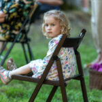 Dziewczynka w sukience w kwiaty siedzi na krześle na trawie. Obok widoczne są inne osoby i kosz piknikowy. Tło jest rozmyte.