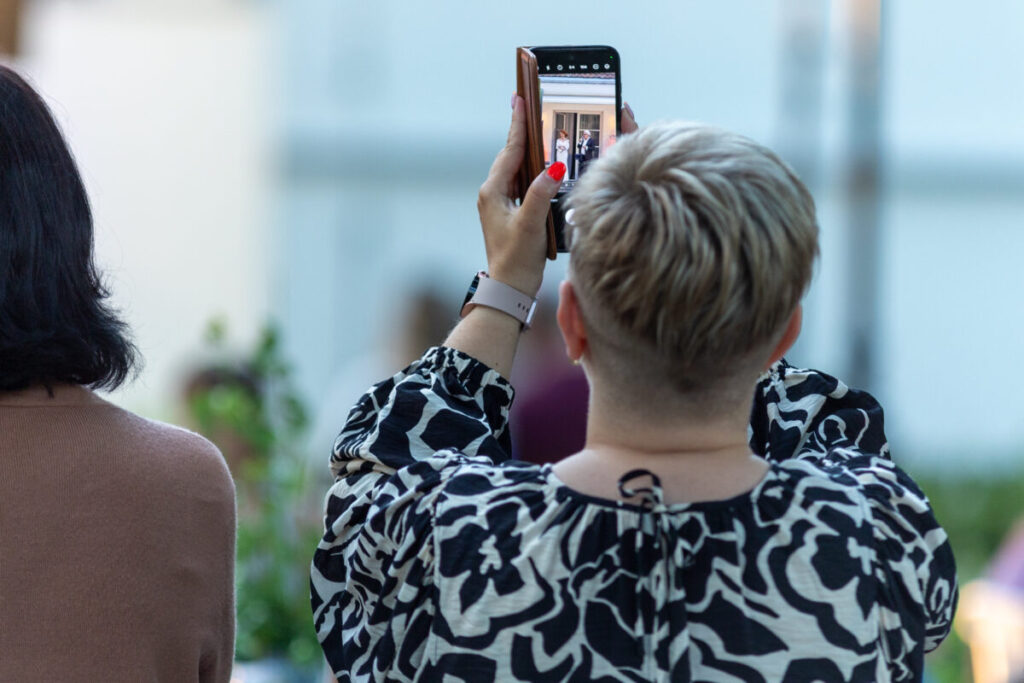 Osoba z krótkimi, blond włosami robi zdjęcie telefonem. Ubrana w czarno-białą bluzkę, na tle rozmytych roślin i innych osób. Jej paznokcie są pomalowane na czerwono.