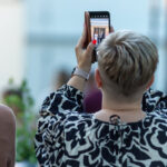 Osoba z krótkimi, blond włosami robi zdjęcie telefonem. Ubrana w czarno-białą bluzkę, na tle rozmytych roślin i innych osób. Jej paznokcie są pomalowane na czerwono.