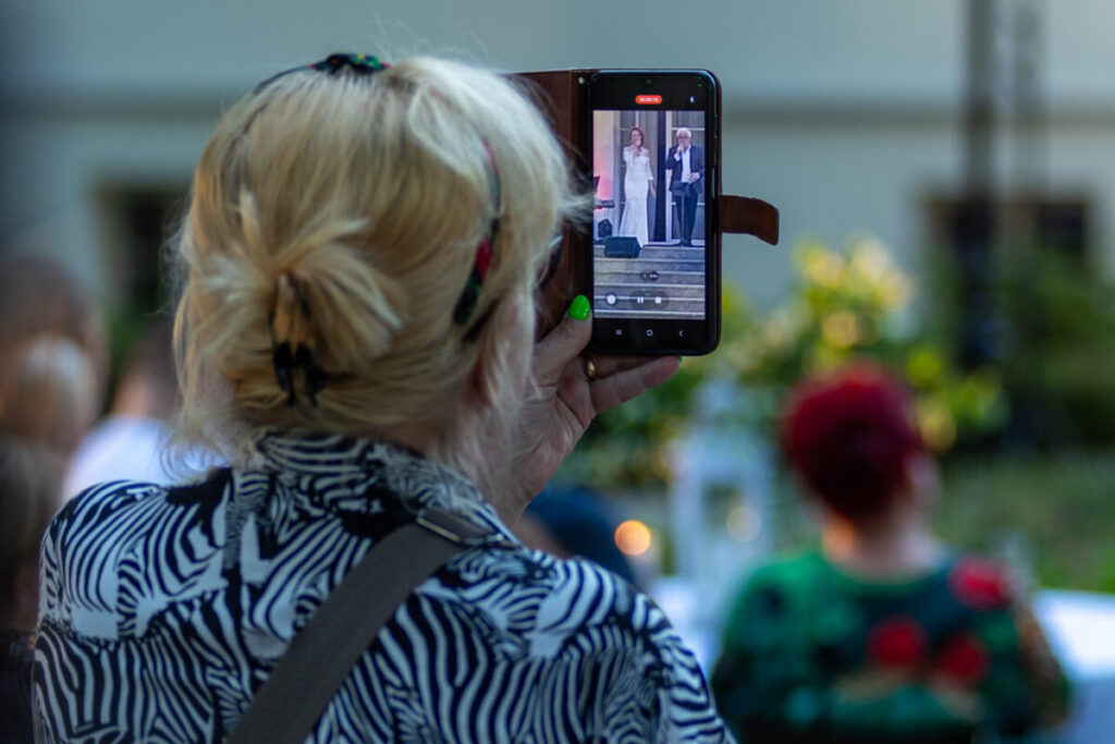 Osoba nagrywa występ kobiety i mężczyzny na scenie za pomocą smartfona. Tło jest rozmyte, dominują zielone i jasne barwy. Widać zebranych ludzi na wydarzeniu plenerowym.