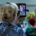 Osoba nagrywa występ kobiety i mężczyzny na scenie za pomocą smartfona. Tło jest rozmyte, dominują zielone i jasne barwy. Widać zebranych ludzi na wydarzeniu plenerowym.