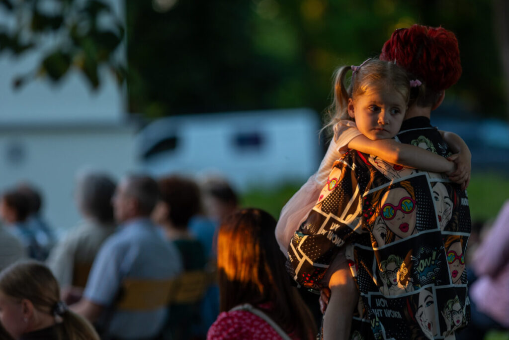 Na zdjęciu kobieta z czerwonymi włosami trzyma na rękach dziewczynkę o blond włosach. Kobieta ma kolorową koszulę z motywem komiksowym. W tle rozmyta grupa ludzi siedząca na krzesłach.
