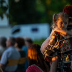 Na zdjęciu kobieta z czerwonymi włosami trzyma na rękach dziewczynkę o blond włosach. Kobieta ma kolorową koszulę z motywem komiksowym. W tle rozmyta grupa ludzi siedząca na krzesłach.