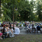 Na zdjęciu widoczna jest grupa osób siedzących na zewnątrz w parku, otoczona zielenią drzew. W tle widać brukowaną ścieżkę i latarnie. Uczestnicy wyglądają, jakby brali udział w wydarzeniu lub spotkaniu.
