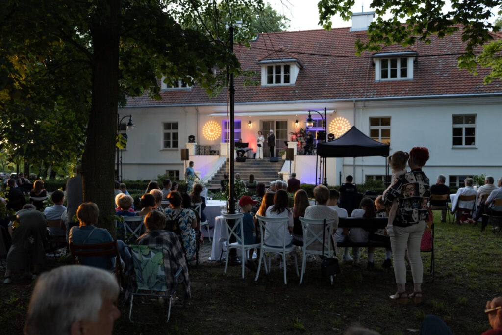 Plenerowy koncert przed białym budynkiem z czerwonym dachem. Publiczność siedzi na krzesłach, scena oświetlona żółtymi i fioletowymi reflektorami. W tle drzewa dodają atmosfery.