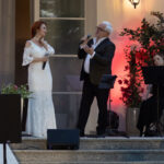 Na podeście stoi kobieta w białej sukni i mężczyzna z mikrofonem. Obok siedzi osoba grająca na flecie. Tło oświetlone na czerwono.