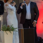 Dwoje ludzi śpiewa na scenie. Kobieta w białej sukni, mężczyzna w czarnej marynarce, tło z czerwonym światłem. Kwiaty w doniczce.