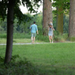 Na zdjęciu widać parę spacerującą z dziećmi po ścieżce w parku. Wokół są wysokie drzewa, zielona trawa i krzewy. Kobieta niesie dziecko na ramionach, a mężczyzna idzie obok.