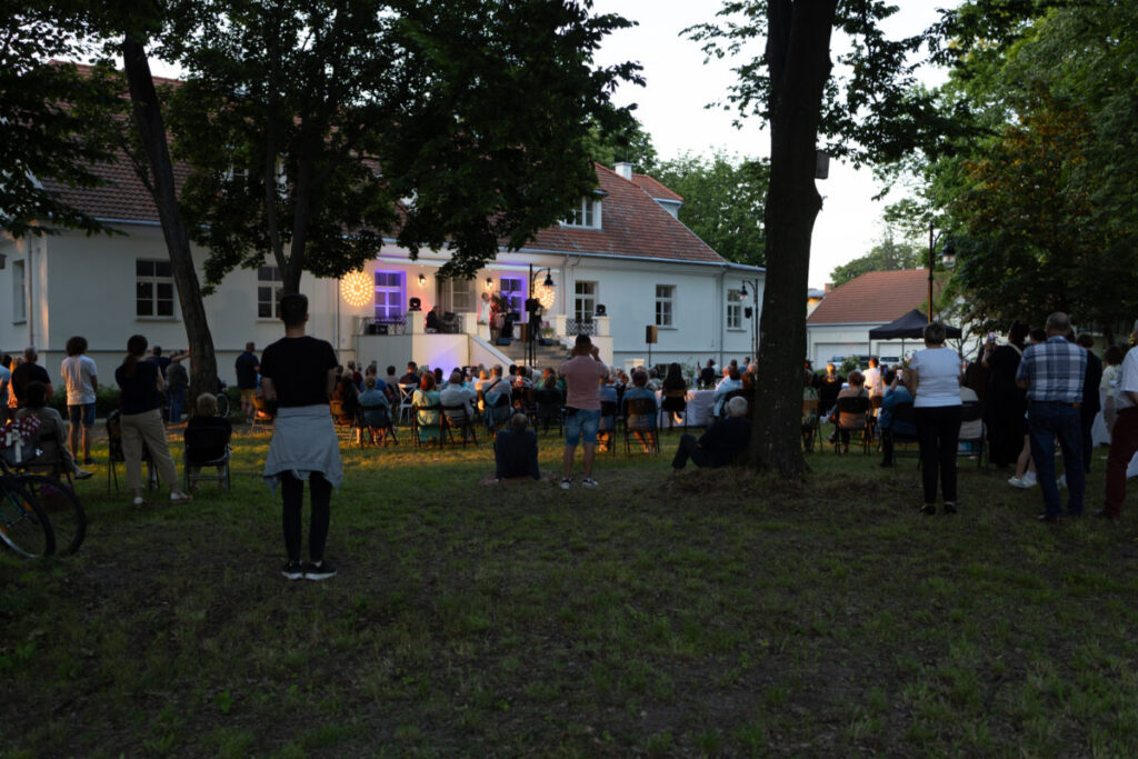 Tłum stoi na koncercie przed białym budynkiem oświetlonym na fioletowo. W tle drzewa i trawniki. Wieczorna atmosfera.