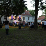 Tłum stoi na koncercie przed białym budynkiem oświetlonym na fioletowo. W tle drzewa i trawniki. Wieczorna atmosfera.